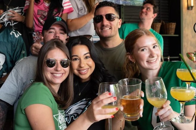 A group of white people hold pints of beers and martini glasses with mimosas and smile. They are wearing Eagles gear at Figo.