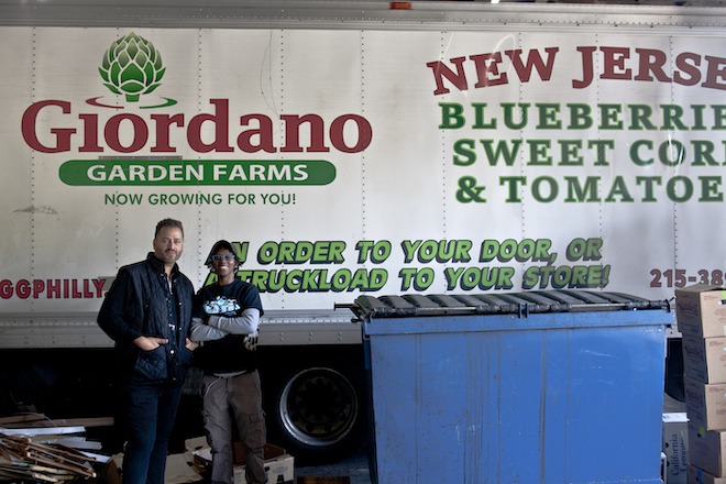 Two people stand in front of the side of a Giordano's Garden Grocery truck. Beside them is a blue dumpster.