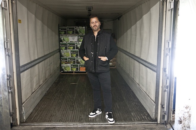 A white man wearing all black stand inside a delivery truck. Behind him are cartons of food.