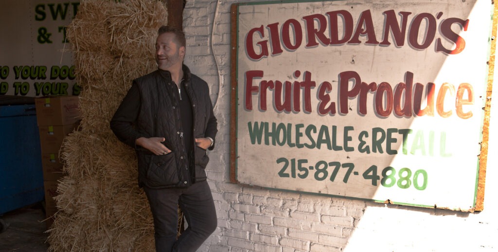 Marcello Giordano, a middle-aged white man with dark hair, stubble, wearing all black, stands with his hands in his pockets leans against a painted brick wall next to an old sign that says Giordano's Fruit & Produce Wholesale.