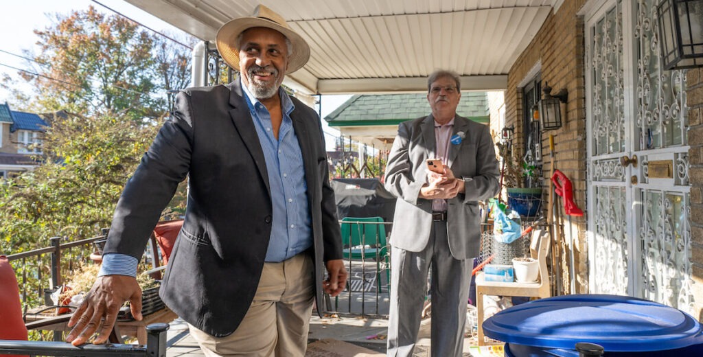 Maurice Sampson (left), an African American man with a wide-brim hat, khaki pants, navy suit jacket and blue collared shirt, stands beside Michael Kleiner, a white man with gray hair and gray mustache wearing a pink shirt, gray suit, buttons on his lapel and holding an iPhone, stand on the porch of a twin home in Mount Airy, Philadelphia.