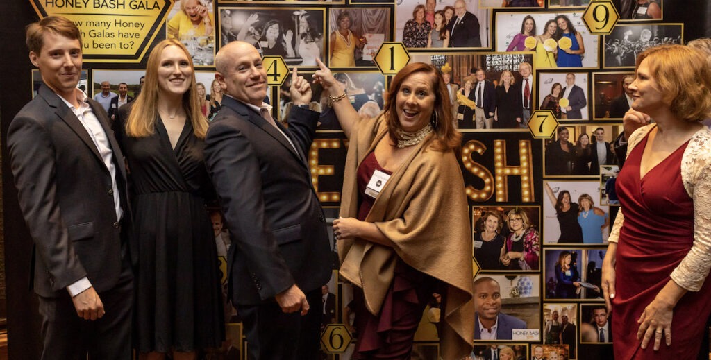 Five White people, two men in back suits and three women in cocktail attire, stand before a wall of photos at the Honey Bash, a fundraiser for brain aneurysm research.