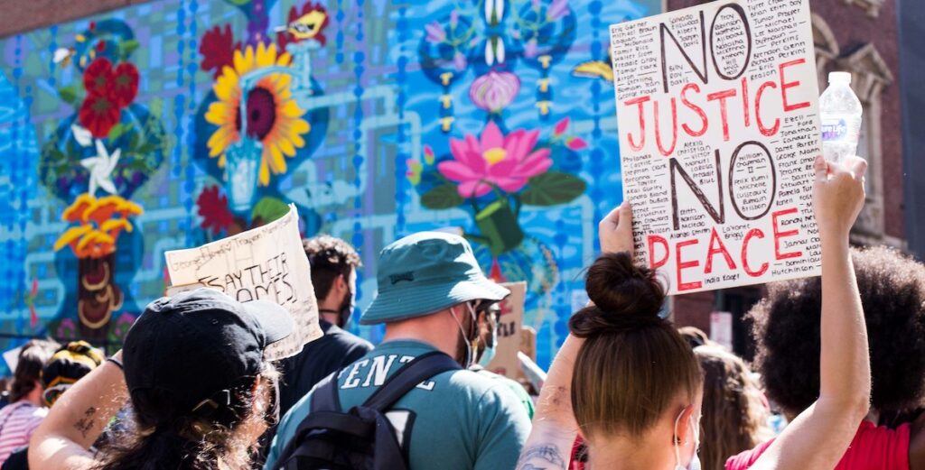 Protesters face a mural wall of flowers. Some hold signs that say "SAY THEIR NAMES" and "NO JUSTICE NO PEACE."