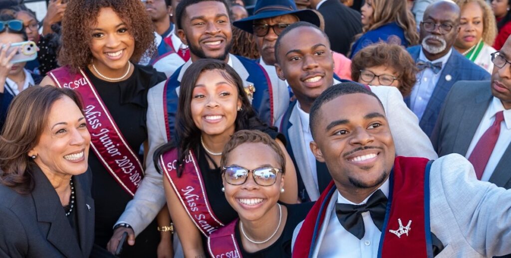 Vice President Kamala Harris poses with African American students from South Carolina State University, who are dressed in formal attire and wearing sashes.