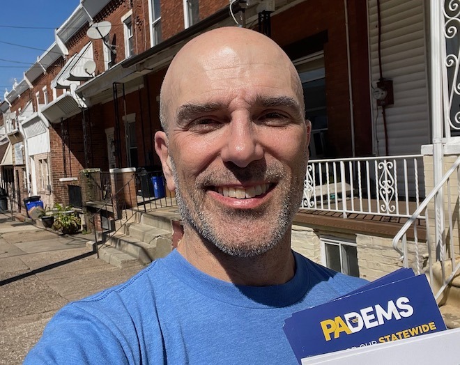 Khris Lewin, a white man with a bald head and blue shirt, stands before a row of homes holding Harris-Walz campaign materials while canvassing in Philadelphia.