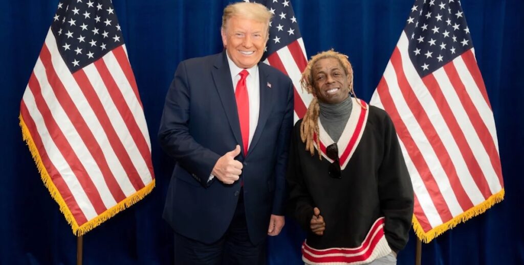 Donald Trup, a White man with combed over blonde hair wearing a large dark suit and long red tie and white shirt, smiles and gives the thumbs-up sign while standing next to Lil Wayne, a smaller Black man with long blonde locs, face tattoos, a grey turtleneck and red, white and blue v-neck sweater. Behind them are three U.S. flags.