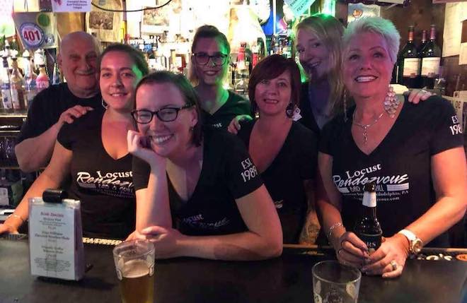 A group of white women and one man, all wearing black t-shirts that say "Locust Rendevous" stand behind a bar and smile.