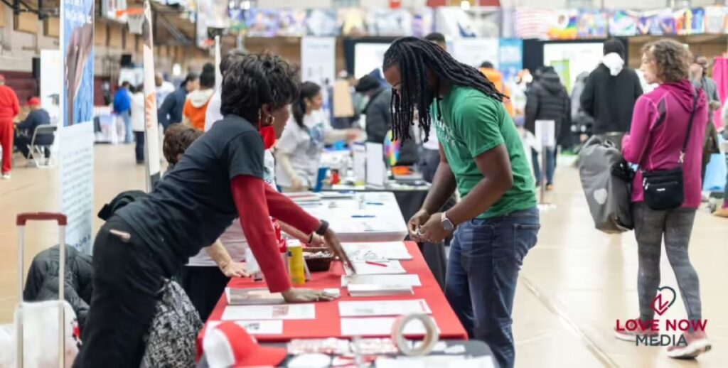 A Black woman in a black t-shirt with red long sleeves and black pants points to political educational materials on a table in at an event as a young Black man with dreadlocks in a green t-shirt and jeans stands across the table from her reading