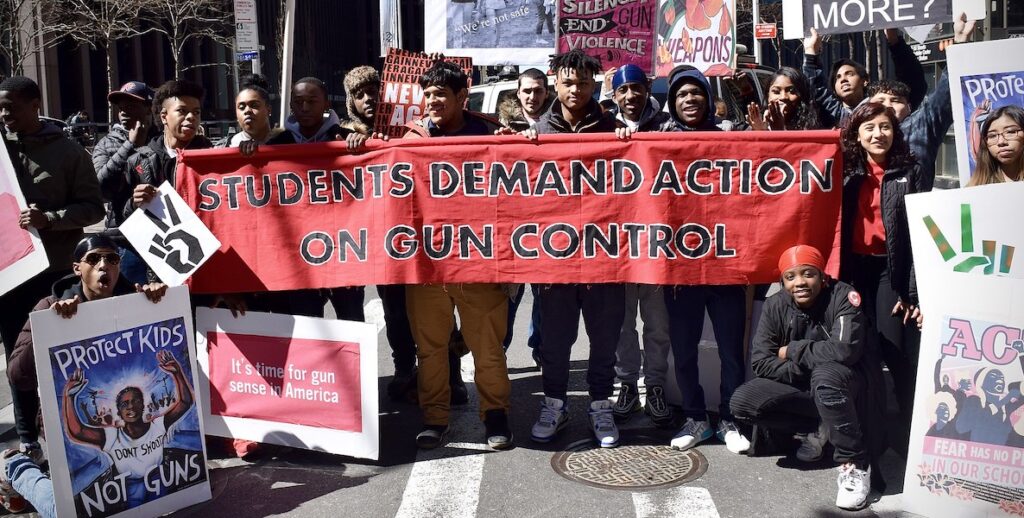 A group of students stand in a street holding a large red sign that says STUDENTS DEMAND ACTION ON GUN CONTROL.