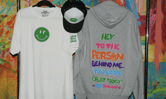 Hanging against a multicolor painted wall, Project Spread Smiles' white t-shirt with a green smiley face with a semicolon for eyes, black and white trucker hat with the same logo, and the back of a hoodie with the multi-color bubble letter message: "HEY. TO THE PERSON BEHIND ME. YOU LOOK GREAT TODAY! KEEP SMILING."