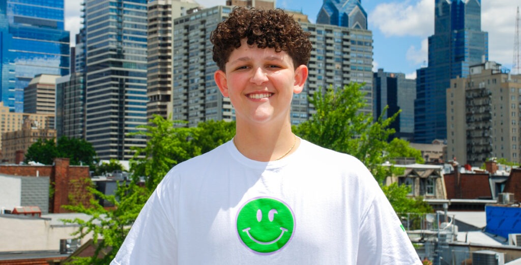 Sammy Grossman, a White teenage boy with short curly brown hair and brown eyes, stands in front of the Center City Philadelphia skyline on a clear day. He is wearing a white t-shirt with a neon green smiley face on the center; the face's eyes are made of a semicolon, a symbol of mental health awareness and support.