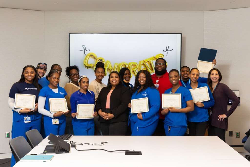 Skills Initiative cohort at Children's Hospital of Philadelphia in 2023. A group of mostly African American women in blue scrubs stand in front of a balloon sign that says "CONGRATS" pinned to a white board. The people in the photo are holding what appears to be certificates. A white table is in front of them.