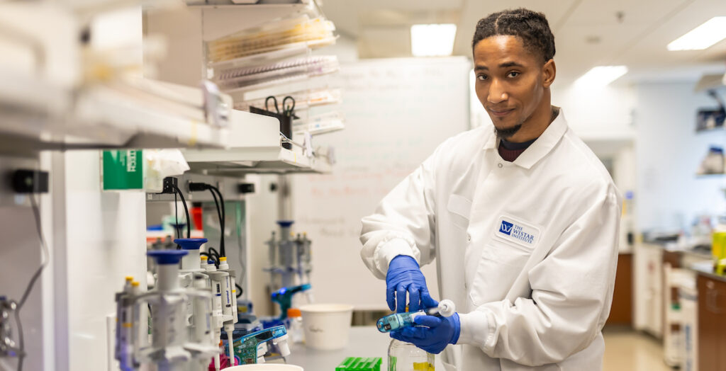 A Skills Initiative worker in a lab at the Wistar Institute. An African American man in a white lab coat and blue plastic gloves holds a piece of lab equipment in a lab setting.