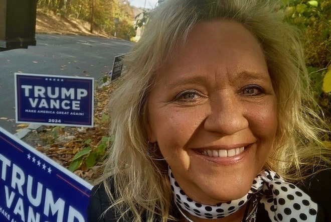 Steffi Behrends, a White woman with bleached blond bobbed hair and a dark tan, stands outside among fall leaves and Trump Vance signs. She moved from Germany to work for the Trump-Vance campaign.