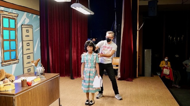 Ted Passon, a white man with grey hair, glasses, a dark mask over his nose and mouth, wearing a white t-shirt with lettering, black pants and sneakers, stands on a movie set behind a girl in a black wig wearing a plaid dress, black and white shoes, and carrying a white script. They are on the set for Patrice: The Movie, on a stage with a desk and an illustrated filing cabinet and window.