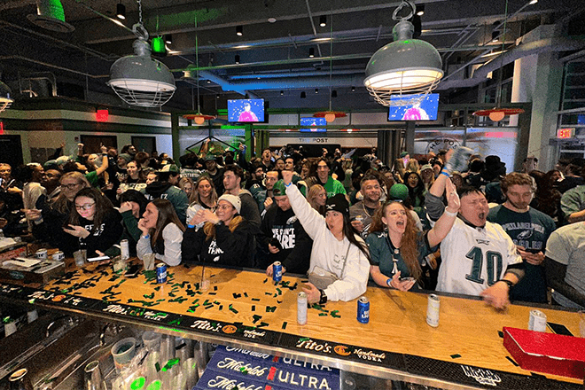 A large group of people wearing Philadelphia Eagles gear crowds into a bar. Some lean on a bar.