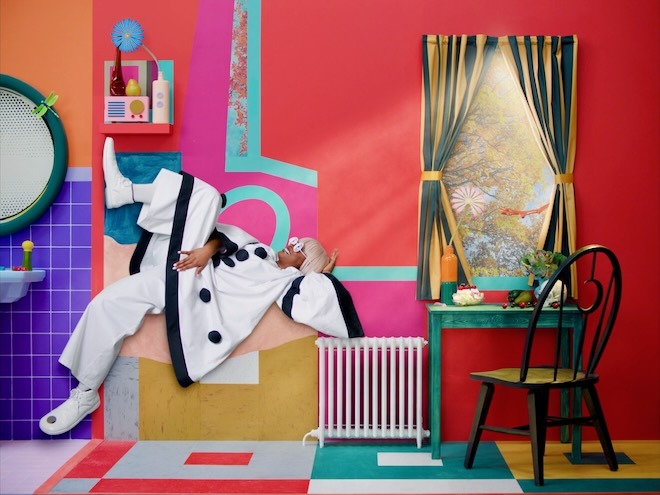 A still from Alex Da Corte's music video for "Shower Song" by Tierra Whack, depicts Whack, a Black woman with a white bob hair style, wearing a white with black trim clown costume, reclining with one leg up and one leg down on a yellow box-like object against a colorful backdrop that includes a green desk and chair and window looking out toward a tree, framed by striped curtains. Beyond Whack's feet is a sliver of a bathroom, with purple square tile, orange paint, and a round mirror.