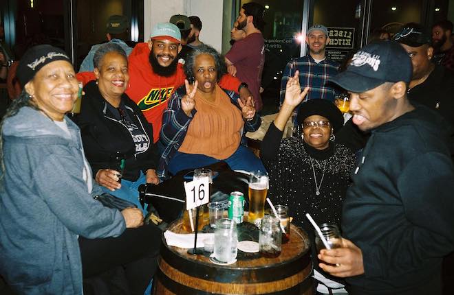 A crowd of mostly African American people sit around a barrel table at Two Locals Brewing in West Philadelphia. The brewery is Black-owned and offers special during Eagles games.