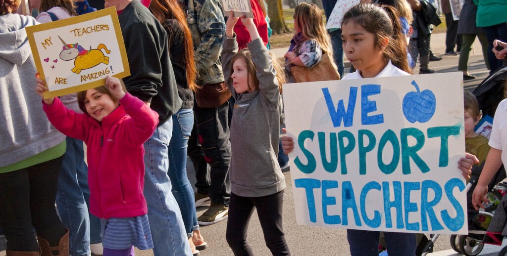 Children hold signs in support of teachers while walking along a street. One sign says "We Support Teachers!"