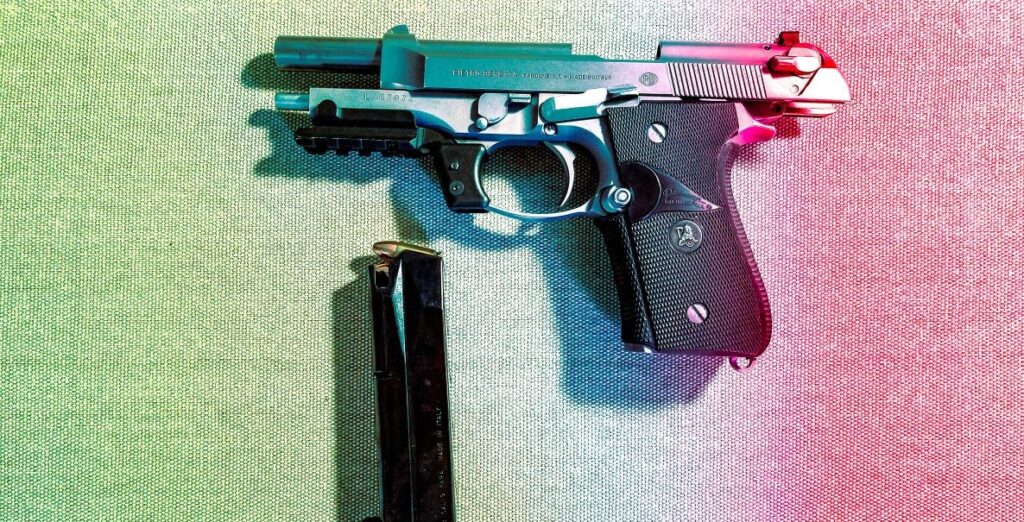 A black and silver semi automatic handgun with a clip beside it sits on a pale rainbow background