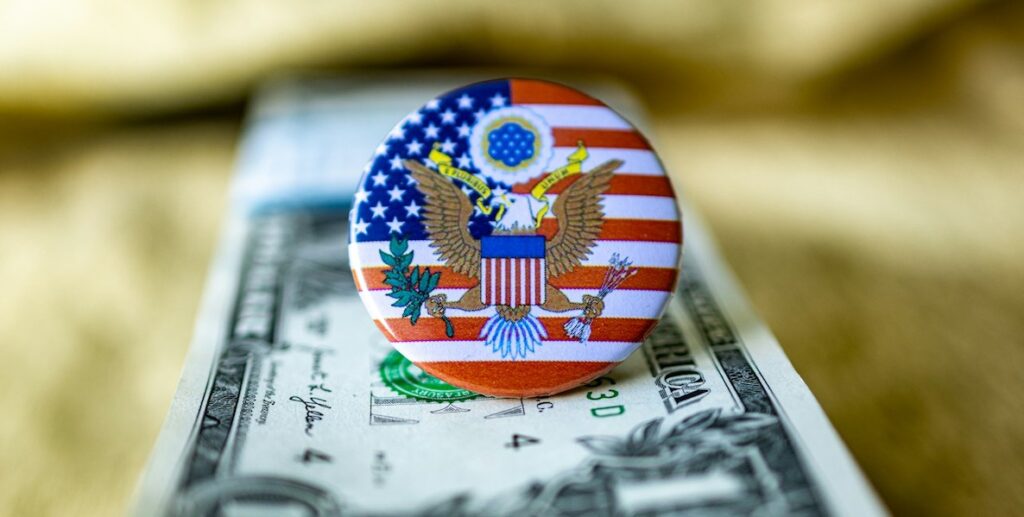 Atop a stack of one U.S. dollar bills is a pin displaying the presidential seal.