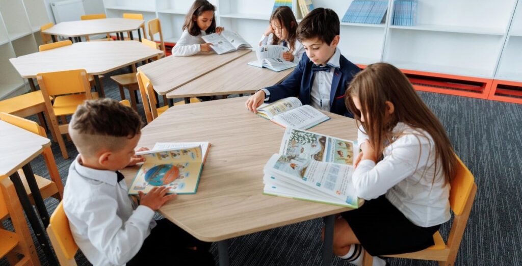 Five elementary school students sit around wooden tables in a bright modern classroom with reading texts open before them