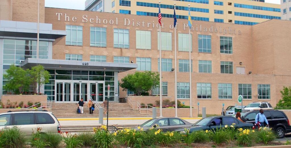 The School District of Philadelphia Building at 400 N. Broad Street. Cars are parked outside, and a street divider contains yellow flowers. People walk down the steps of the blocky building.