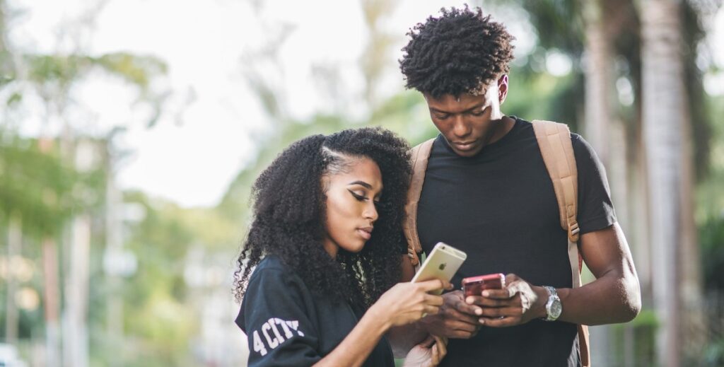 Two African American people, a woman with long, full hair and a black top holding and looking at an iPhone, and a taller man with short twists wearing a black t-shirt, backpack and silver watch, also looking at a phone that he's holding. Behind them are trees.