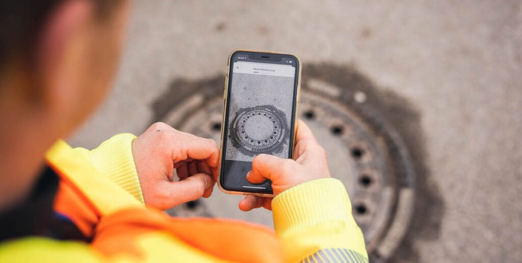 A blurred partial view of the back of a man wearing a yellow safety jacket, holding a phone and taking a photo of a manhole for vialytics, an ai company that tracks potholes and other roadway problems.