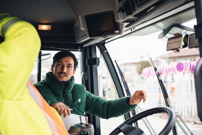 Two people inside a vehicle. One is seated behind a steering wheel; that person's face is not visible, but they appear to be wearing bright safety gear. The other person is facing the seated person, wearing a green puffer jacket, with short dark hair and a mustache. A small phone mounted to the windshield displays the vialytics app, software that tracks and maps potholes and other roadway problems.