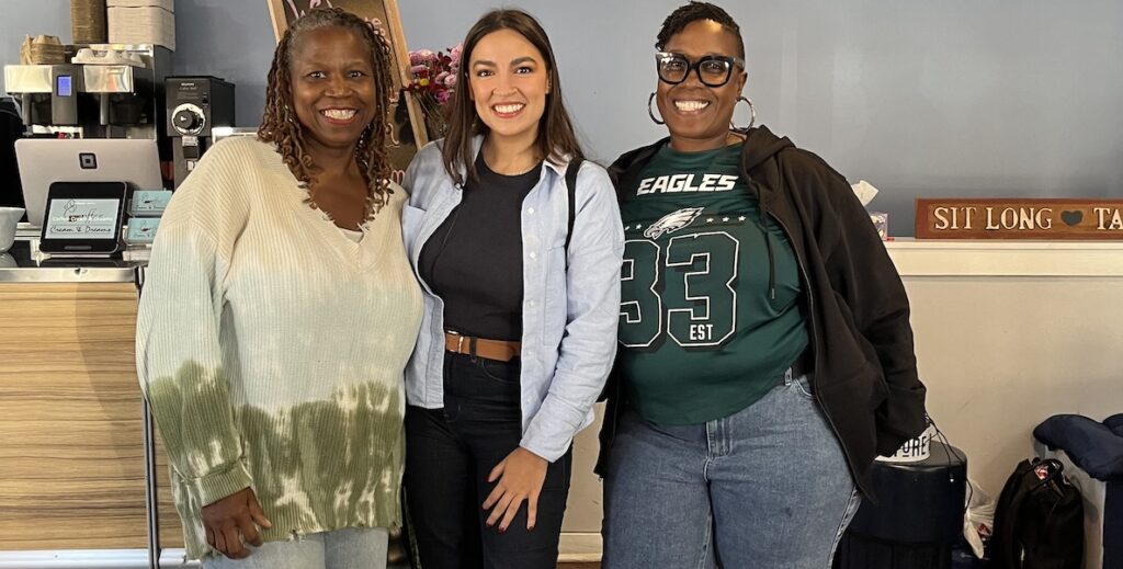 Coffee, Cream & Dreams co-owner Stephanie Ford, a Black woman with braids wearing a tie-dye v-neck sweater, stands alongside, with her arm behind U.S. Representative Alexandria Ocasio-Cortez, a Latina woman with long dark brown hair wearing a denim button-down shirt opened over a black t-shirt and black years, next to cafe co-owner Sonja West, a Black woman in an Eagles 33 shirt, dark hoodie and jeans, glasses and large hoop earrings with short hair at the Fairmount cafe and co-working space.