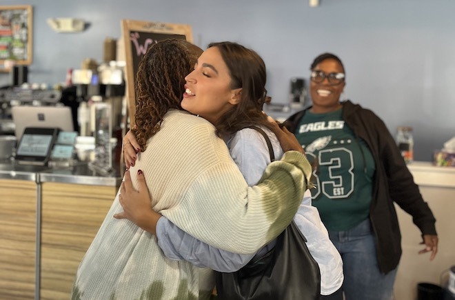 Coffee, Cream & Dreams co-owner Stephanie Ford, a Black woman with braids wearing a tie-dye sweater, embraces U.S. Representative Alexandria Ocasio-Cortez, a Latina woman with long dark brown hair holding a large black leather bag and wearing a denim shirt, while cafe co-owner Sonja West, a Black woman in an Eagles 33 shirt, dark hoodie and jeans, glasses with short hair smiles in the background of the shot at their Fairmount cafe and co-working space.