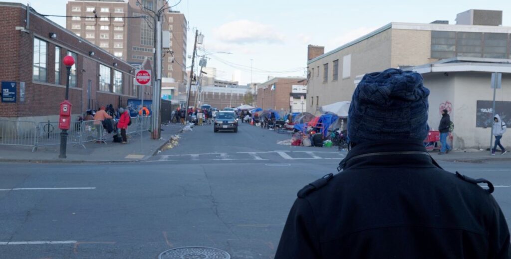 A man in a black jacket and a blue winter cap stands across from an intersection. The opposite street is lined with tents where homeless people have made their shelter. Nondescript brick buildings are to the left and right of the street