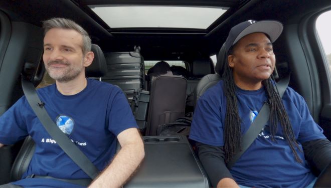 Two men in matching blue t shirts are sitting in a car with equipment piled up behind them in the cargo area. The man on the left is white with salt and pepper hair, beard, and mustache, while the man on the right is Black with long black dreadlocks and a black baseball cap.