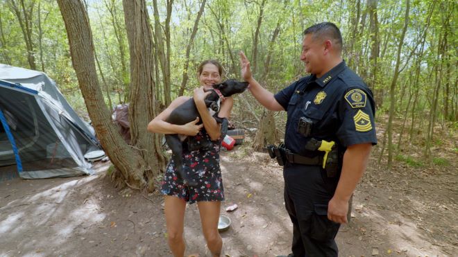 A sunny day in the woods, a large tent can be seen to the left, in the foreground is a woman in a short black flowered jumper holding a black and white terrier mix and to the right of her stands a male police officer in a black uniform petting the doggo.