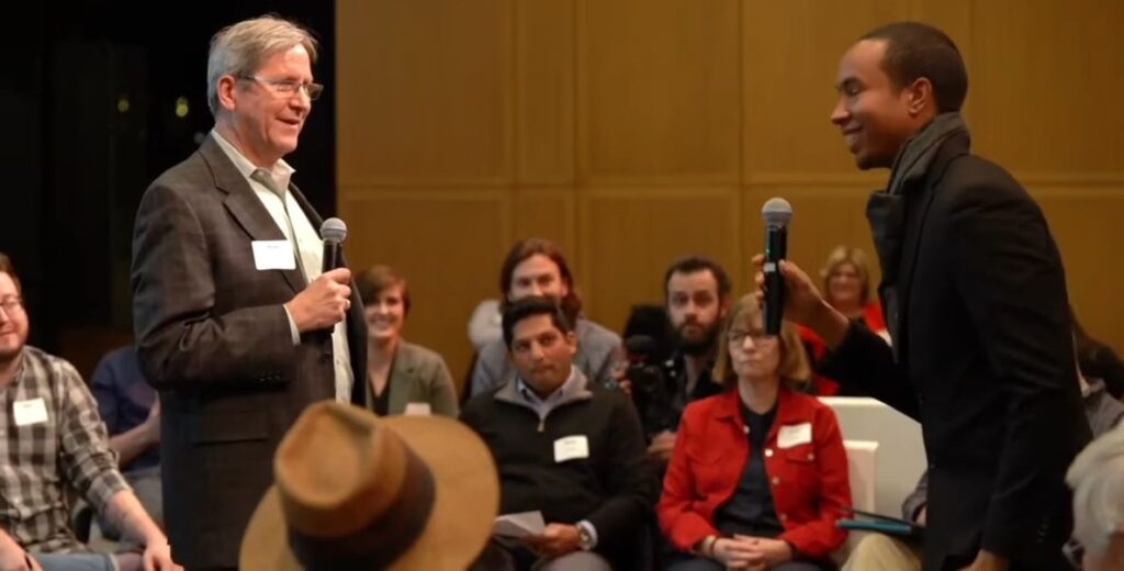 In a still from a documentary film, an older white man in a grey suit jacket stands a few feet in front of a young Black man in a long-sleeved shirt, each holding a microphone. Behind them is a room of seated people with nametags