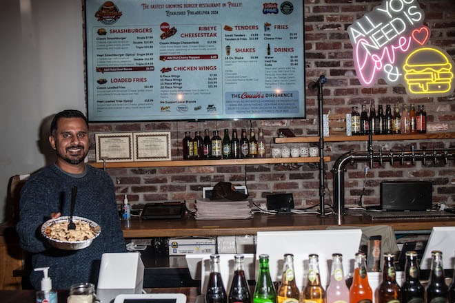 Shahezad Contractor, an Indian American man with short black hair, mustache and close beard wearing a grey knit sweater, holds an aluminum foil container of food while standing in the Poplar Street Cousin's Burger, against a backdrop of exposed brick, a wall menu and bottles of Hank's soda.