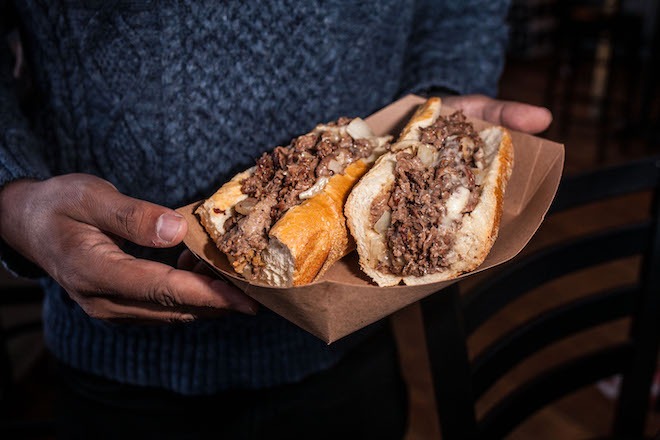 Brown hands hold a paper container with a cheesesteak. The holder wears a grey knit sweater.