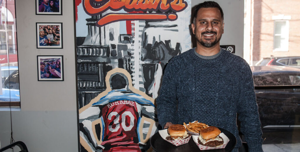 Shahezad Contractor, an Indian American man with black and grey short hair, a black beard and mustache, holds a tray containing paper containers of cheeseburgers and fries in the Poplar Street Cousin's Burger. Behind him is a mural depicting the back of 76ers player Kuramaz, the word "Cousin's," and framed photos. Also: windows.