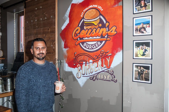 Shahezad Contractor, an Indian American man with black hair, mustache and short beard wearing a grey sweater stands next to a wall mural that says "Cousin's Burger" and "city of BURGERLY LOVE" with an illustration of a cheeseburger, alongside some framed photos in the Poplar Street Cousin's Burger.