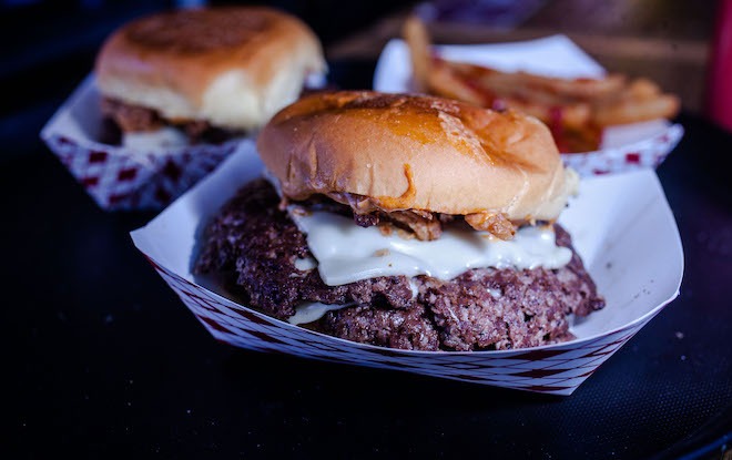 Two oversized cheeseburgers on smaller buns and in the background, french fries with ketchup, all in paper containers.