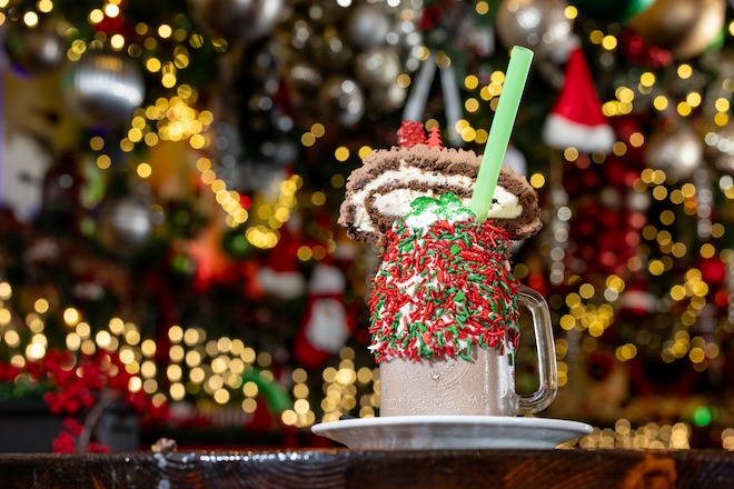 A "Fab-YULE-Ous" milkshake at Craftsman Row holiday bar is a glass mug covered on the outside with sprinkles with a slice of yule log cake and straw, backdropped by many decorations. 