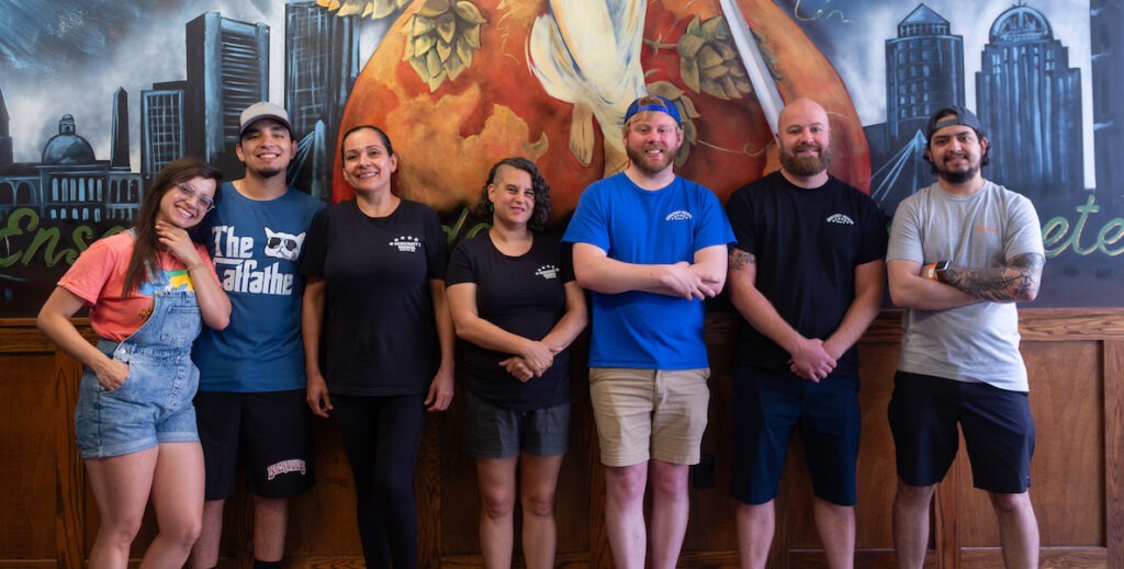 Seven people in casual dress, some in t-shirts with logos that say "Democracy Brewing" stand against an interior mural. They are mostly white and Latino.