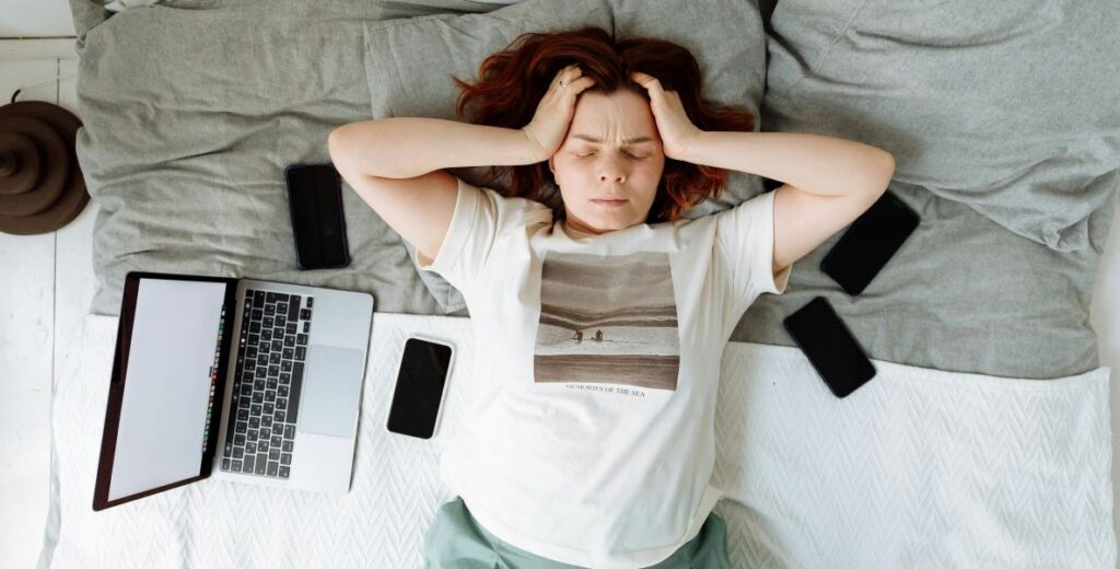 A red-haired white woman in a white graphic t-shirt holds her head in her hands at the temples and squeezes her eyes closed as she lays on a bed with gray pillows and a white blanket. Her laptop is on the left on her bed and she is surrounded by four smartphones