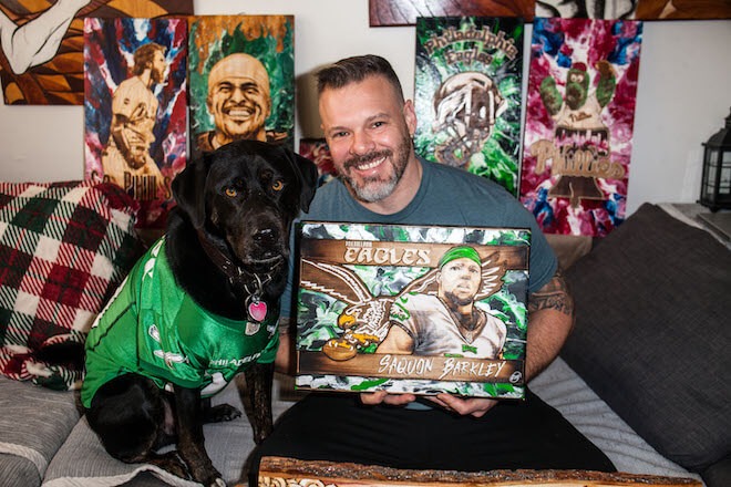 Jon Metz, a White man with short dark hair, a grey-and-white beard, grey t-shirt sits beside his dog, who is wearing a Kelly green Eagles jersey. Metz is holding a wood burned image of Saquon Barkely with the Eagles logo. Behind him stand other athlete-inspired works.