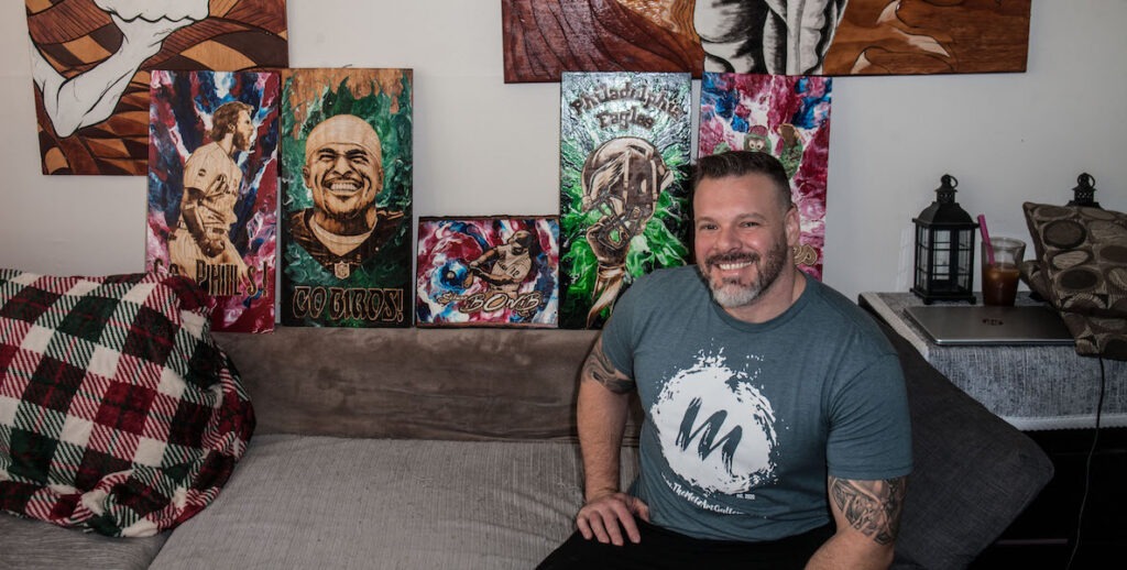 Jon Metz, a White man with short dark hair, a grey-and-white beard, grey t-shirt and tattooed arms, sits in front of his burned wood works, including portraits of Bryce Harper and Jalen Hurts.