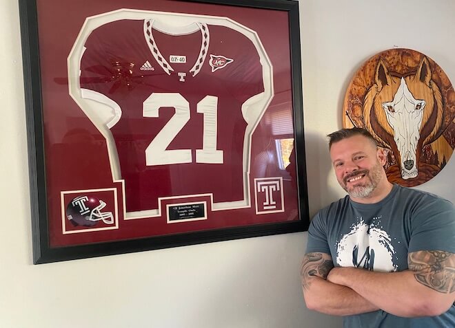 A large framed Temple football jersey hangs on a white wall beside a wood burned portrait of a wolf face. Standing between these items is Jon Metz, a White man with short dark hair, a grey-and-white beard, grey t-shirt and tattooed folded arms.