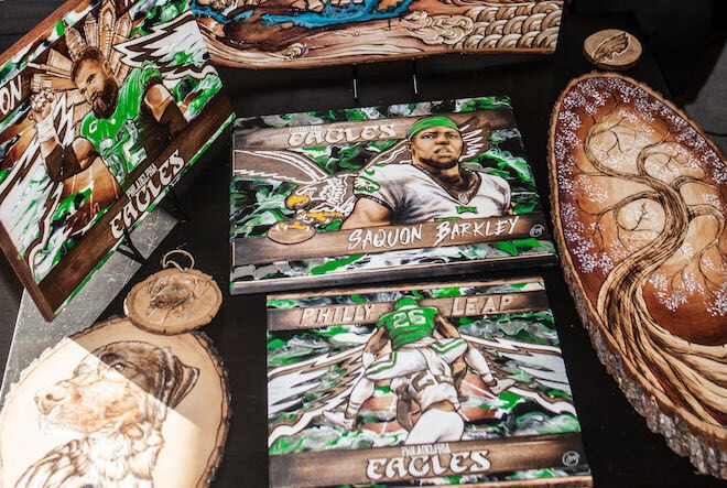 Wood burned works include portraits of Eagles players Jason Kelce and Saquon Barkley, an illustration of Barkley leaping over a Jets player, and two dog portraits.