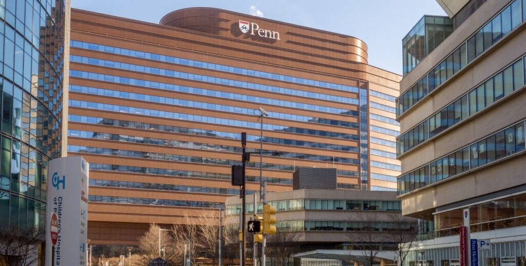 The copper-toned and striped with windows Penn Medicine Pavilion located at the Hospital of the University of Pennsylvania is set against a blue sky with the sun behind surrounding buildings