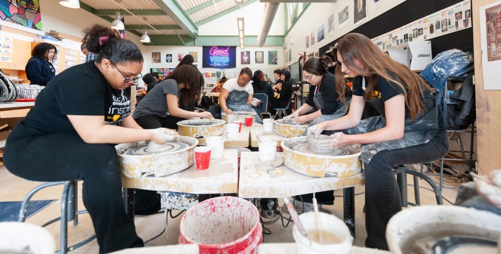 High school students in an art classroom sit in chairs facing each other, leaning over a table filled with potters' wheels, working on ceramics as part of an after-school time school enrichment program of the School District of Philadelphia.